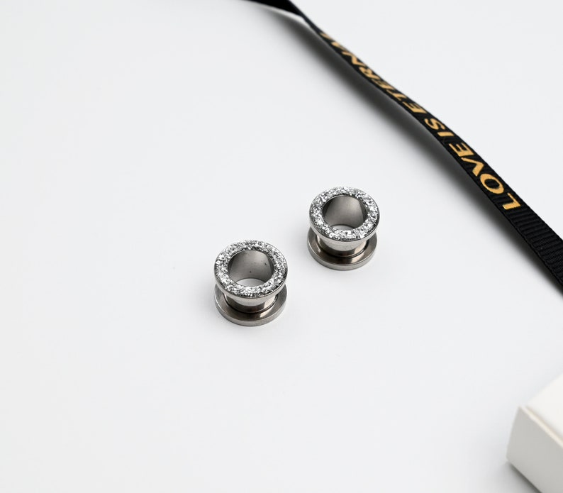 Puede incluir: Dos expansores de oreja plateados con un anillo de peque&ntilde;os cristales transparentes en el borde exterior. Los expansores son circulares con un dise&ntilde;o acampanado. Una cinta negra con texto dorado que dice "LOVE IS ETERNAL" en el fondo.