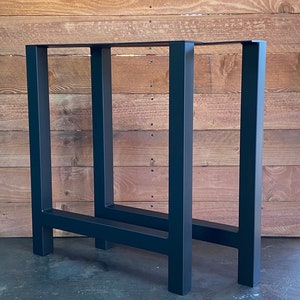 May include: Two black metal table legs with a rectangular frame. The legs are made of sturdy metal and have a black powder coat finish.