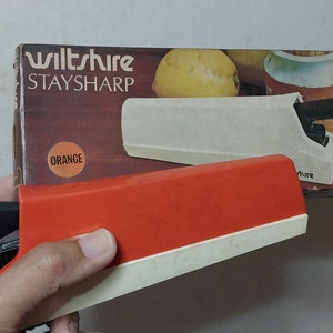 Puede incluir: Un afilador de cuchillos Wiltshire StaySharp naranja y blanco con mango negro. El producto está en su caja original, que presenta el nombre de la marca y una imagen del cuchillo. La caja es naranja y marrón.