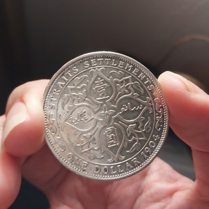May include: A silver Straits Settlements 1904 One Dollar coin. The coin has intricate text and patterns around the edges and in the center. The coin is held between two fingers.