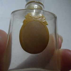 May include: A small, clear glass perfume bottle with a rounded top and a clear stopper. The bottle has a light brown oval label with the word "Christian" visible. The label is adorned with a bow design. The bottle is held by a person.