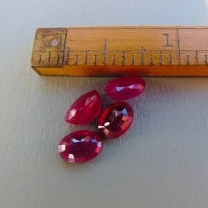 May include: Four oval-shaped, deep red gemstones. The gemstones are arranged in a cluster on a grey surface.