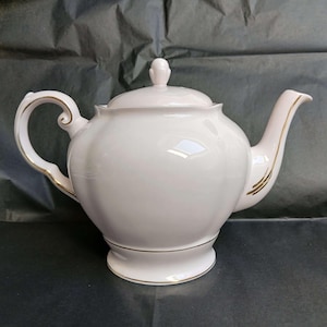 May include: A white porcelain teapot with a rounded, fluted body and a matching lid featuring a small finial. The teapot has a curved handle and a spout, both accented with thin gold trim, and a gold band around the base and the spout opening.