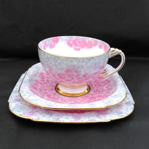 Può includere: Un servizio da tè vintage composto da tazza, piattino e piattino. Il set presenta un motivo floreale rosa e bianco con bordi dorati. La tazza, il piattino e il piattino sono disposti su uno sfondo nero.