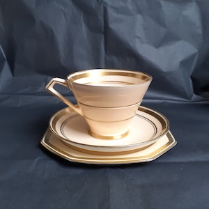 May include: A cream-colored teacup and saucer set with gold trim. The cup has a flared shape and the saucer is octagonal.