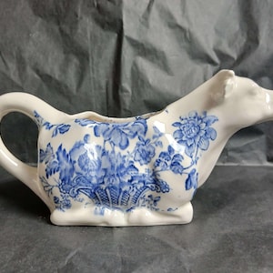 May include: A white ceramic creamer shaped like a cow, featuring a blue floral pattern across its body. The handle is formed by the cow's tail, and the spout is located at the head. The design includes detailed blue flowers and foliage.