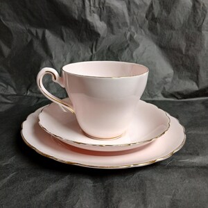May include: A pale pink teacup, saucer, and side plate set. The set features a gold trim. The teacup has a curved handle. The saucer and side plate have a scalloped edge. A classic tea set.