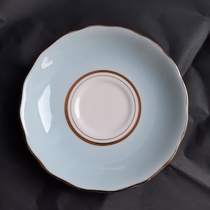 May include: A light blue saucer with a scalloped edge and a gold rim. The center of the saucer is white with a gold border. The saucer is likely part of a tea set.