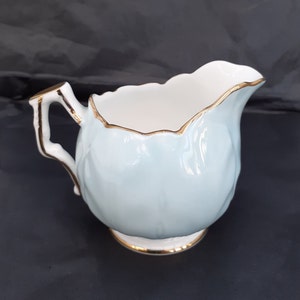 May include: A light blue creamer with a gold trim and a ruffled edge. The creamer has a white interior and a gold handle.
