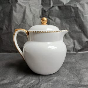May include: A white porcelain creamer with a gold handle and trim. The creamer has a lid with a gold knob. The design is simple and elegant, suitable for serving cream or milk. The creamer is approximately 15 cm tall.