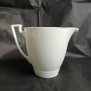 Può includere: Una lattiera in ceramica verde pallido con un design unico del manico. La lattiera ha un beccuccio curvo e un bordo dorato. La base presenta tre scanalature orizzontali. Un pezzo vintage.