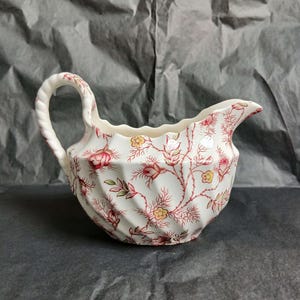 May include: A cream-colored ceramic creamer featuring a swirling, fluted body and a twisted handle. The surface is decorated with a repeating pattern of delicate red floral vines, green leaves, and small yellow blossoms across the entire exterior.