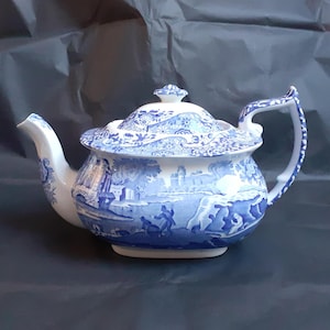 Puede incluir: Una tetera azul y blanca con un diseño floral y una escena de un castillo y un puente. La tetera tiene un cuerpo redondo y un pico curvo.