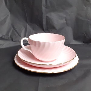 May include: A pink teacup and saucer set with a gold trim. The teacup has a swirling design and the saucer has a scalloped edge.