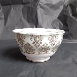 May include: A white ceramic bowl with a gold ornate pattern. The bowl has a scalloped rim and a pedestal base. The gold pattern is a repeating design of swirls and floral elements. The bowl is likely vintage or antique.