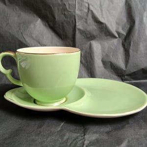 Könnte beinhalten: Eine hellgrüne Teetasse aus Keramik mit passendem Snackteller. Die Tasse verfügt über einen geschwungenen Henkel und einen goldfarbenen Rand. Der Teller hat eine kreisförmige Vertiefung für die Tasse und bietet Platz für kleine Speisen.