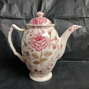 May include: Cream-colored ceramic teapot with a floral pattern in shades of red, pink, and green. The teapot has a rounded body, a curved spout, and a handle. The lid is topped with a small knob. The floral design covers the entire surface.