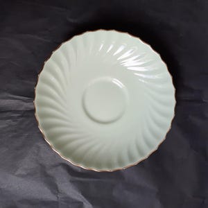 May include: A white saucer with a swirl design and a gold rim. The saucer is made of ceramic and is perfect for serving tea or coffee.