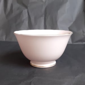 Puede incluir: Un cuenco de cerámica rosa pálido con borde y ribete dorado en la base. El cuenco tiene forma redondeada y se apoya sobre una pequeña base circular. El interior es liso y el exterior es mate.