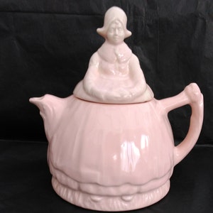 Peut inclure: Une théière en céramique rose avec un couvercle en forme de figurine de fille. La théière a un corps rond et un bec verseur sur le côté. La figurine de fille porte une robe rose et un bonnet blanc.