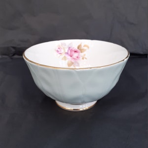 Könnte beinhalten: Eine hellblaue Zuckerdose mit goldfarbenem Rand und einem Blumendesign mit rosa Rosen. Die Schale hat ein geriffeltes Design und einen kleinen Fuß.