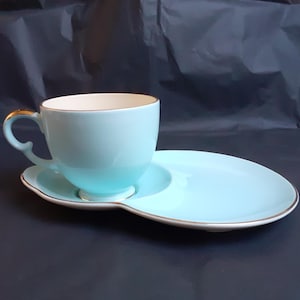Puede incluir: Juego de taza y platillo azul claro. La taza tiene un asa y borde dorados, y el platillo es de forma alargada. El conjunto es de color azul claro liso con ribetes dorados, probablemente de cerámica.
