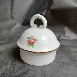 May include: A white ceramic jar with a lid decorated with a rose floral pattern. The lid has a handle and a gold trim. The jar is likely for storing small items or as a decorative piece.