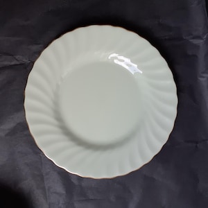 May include: A white ceramic plate with a scalloped edge and a subtle swirl design. The plate has a gold rim.