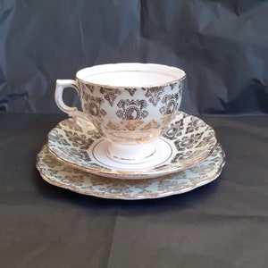 May include: A white teacup and saucer set with a gold and black floral pattern. The teacup has a delicate handle and the saucer has a scalloped edge.