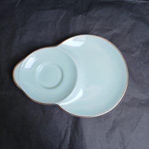 May include: A pale blue ceramic serving dish with a gold rim. The dish has a distinctive shape, featuring a circular indentation on one side, ideal for a cup or small bowl. The dish is set against a dark backdrop.