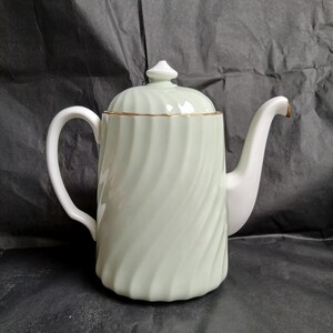 May include: A light green ceramic teapot with a white handle and spout. The teapot has a spiral design and a gold rim. The lid is topped with a small white finial. The teapot is on a dark background.