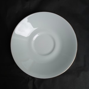 May include: A white ceramic saucer with a gold trim. The saucer features a shallow, circular design with a slightly raised centre. The surface is smooth and reflective, with a glossy finish. The saucer is set against a black background.
