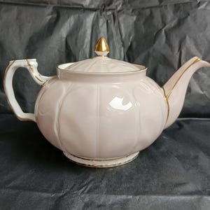 Puede incluir: Una tetera de cerámica rosa claro con detalles dorados. La tetera tiene un cuerpo redondeado con estrías verticales, un pico curvo y un asa a juego. La tapa también es rosa claro con un adorno dorado.