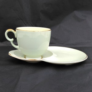 Puede incluir: Un juego de taza y platillo de té de color verde claro con ribete dorado. El platillo tiene una pequeña hendidura para la taza de té.