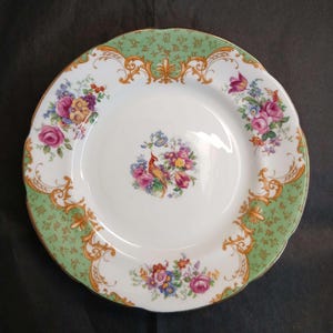 Pode incluir: Um prato redondo de porcelana branca com um ramo de flores central e um pequeno pássaro, rodeado por uma borda larga de cor verde clara com volutas douradas e grupos de flores adicionais em tons de rosa, roxo, amarelo e azul.