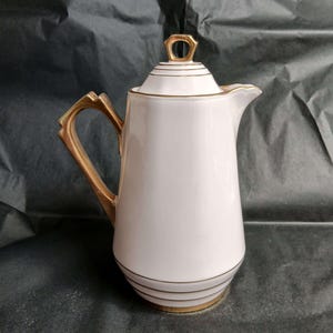 May include: A white porcelain coffee pot featuring a gold-toned handle and a matching gold-toned lid with a geometric finial. The body of the pot is decorated with thin horizontal gold bands near the base and the lid, and it has a spout.