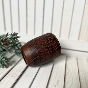May include: A small, brown, barrel-shaped ceramic cup with a floral design carved into the surface. The cup has a dark, textured appearance and sits on a white wooden surface. The cup is likely used for drinking.
