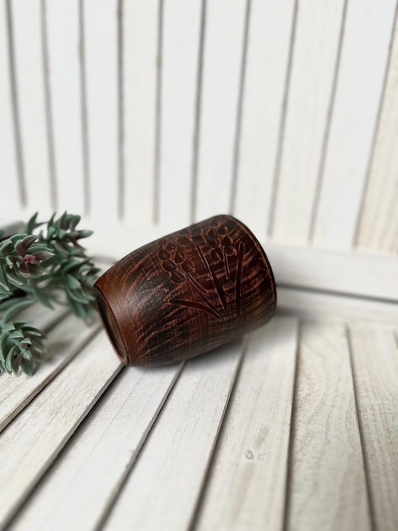 May include: A small, brown, barrel-shaped ceramic cup with a floral design carved into the surface. The cup has a dark, textured appearance and sits on a white wooden surface. The cup is likely used for drinking.