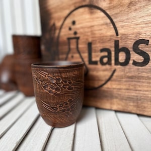 May include: A handmade, brown ceramic cup with a wheat pattern. The cup is cylindrical and sits on a white slatted surface. In the background is a wooden sign with the word "Labs" and a lab flask logo.