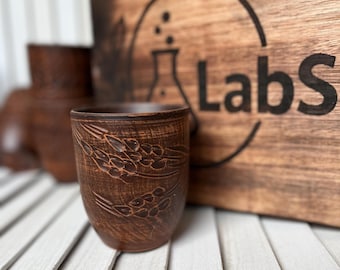 May include: A handmade, brown ceramic cup with a wheat pattern. The cup is cylindrical and sits on a white slatted surface. In the background is a wooden sign with the word "Labs" and a lab flask logo.