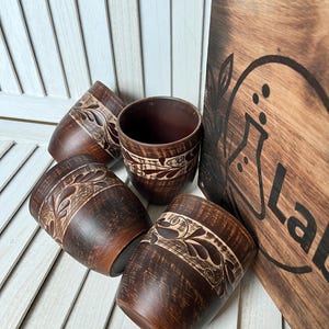 May include: Four brown ceramic cups with a carved floral design and a wooden board with the word "Lab" are displayed. The cups have a dark brown exterior and a lighter brown interior. The wooden board is also brown.