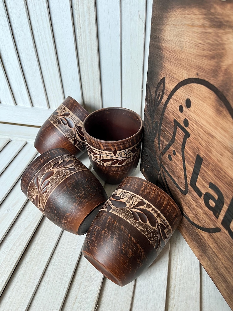 May include: Four brown ceramic cups with a carved floral design and a wooden board with the word "Lab" are displayed. The cups have a dark brown exterior and a lighter brown interior. The wooden board is also brown.