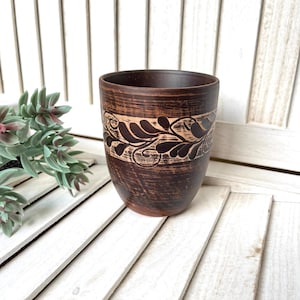 May include: A handcrafted brown ceramic cup with a decorative leaf and vine pattern around the top. The cup has a dark brown exterior and a lighter brown band with the design. The cup is likely used for drinking.