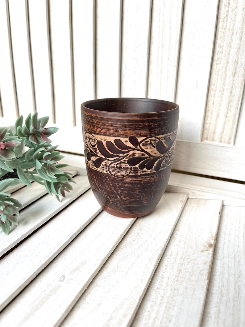 May include: A handcrafted brown ceramic cup with a decorative leaf and vine pattern around the top. The cup has a dark brown exterior and a lighter brown band with the design. The cup is likely used for drinking.