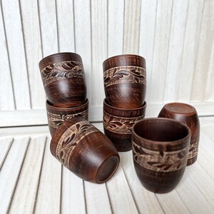 May include: A collection of handmade, dark brown ceramic cups. Each cup features a carved floral design around the top. The cups are arranged on a white wooden surface, showcasing their rustic charm and craftsmanship. The cups are approximately 3 inches tall.