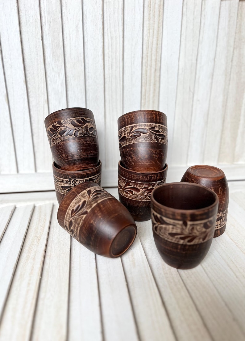 May include: A collection of handmade, dark brown ceramic cups. Each cup features a carved floral design around the top. The cups are arranged on a white wooden surface, showcasing their rustic charm and craftsmanship. The cups are approximately 3 inches tall.