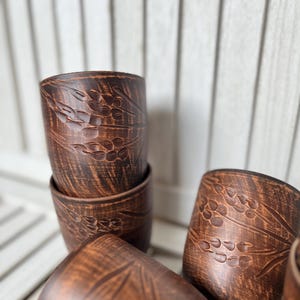 May include: A collection of handmade, brown ceramic cups. Each cup features a detailed, embossed wheat stalk design. The cups are cylindrical and appear to be stacked and arranged on a light-colored wooden surface.