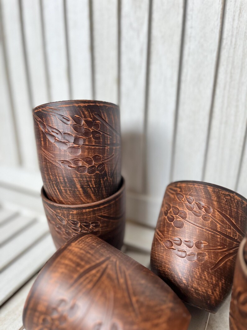 May include: A collection of handmade, brown ceramic cups. Each cup features a detailed, embossed wheat stalk design. The cups are cylindrical and appear to be stacked and arranged on a light-colored wooden surface.