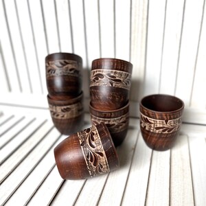May include: A collection of handcrafted, dark brown wooden cups. Each cup features a carved floral design around the top. The cups are arranged on a white slatted surface, showcasing their rustic charm and unique craftsmanship.