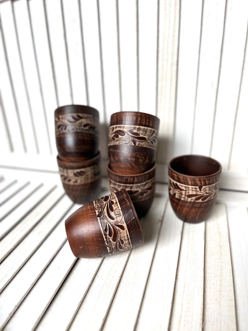 May include: A collection of handcrafted, dark brown wooden cups. Each cup features a carved floral design around the top. The cups are arranged on a white slatted surface, showcasing their rustic charm and unique craftsmanship.